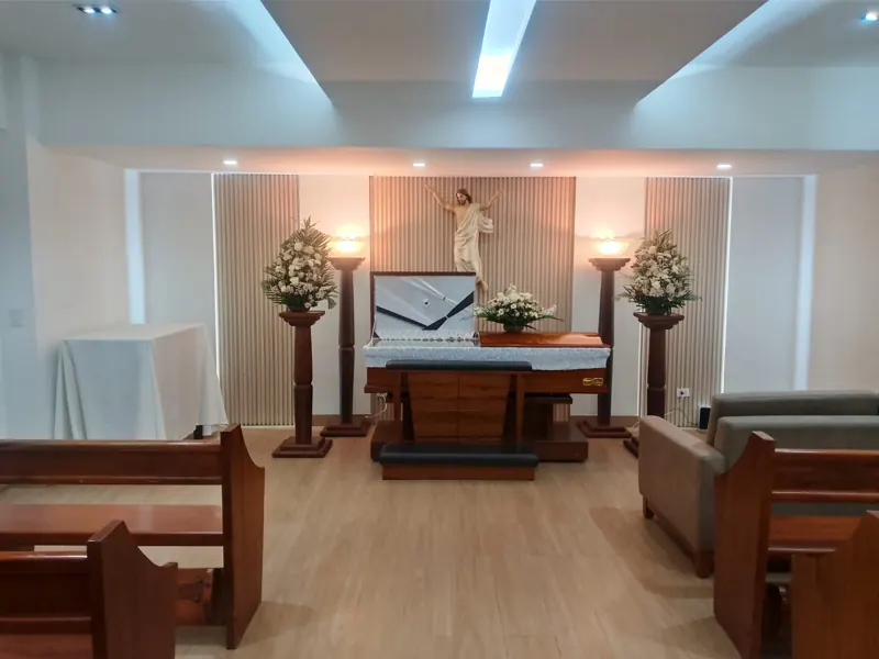 Sala velatoria amplia y luminosa con sofás, arreglos florales y ataúd